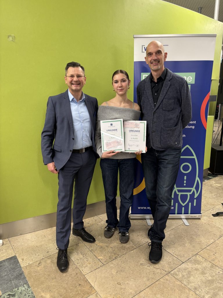 Schülerin präsentiert Urkunden; daneben stehen der Schulleiter und der Lehrer für Wirtschaft und Recht