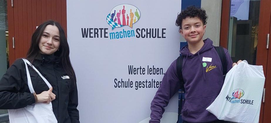 Schülerin und Schüler mit Stoffbeuteln und blauem Koffer vor Plakat der Wertebotschafter