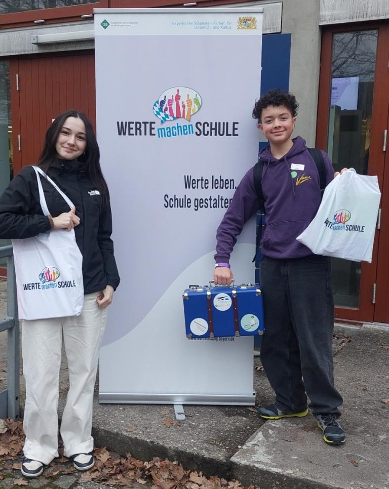Schülerin und Schüler mit Stoffbeuteln und blauem Koffer vor Plakat der Wertebotschafter