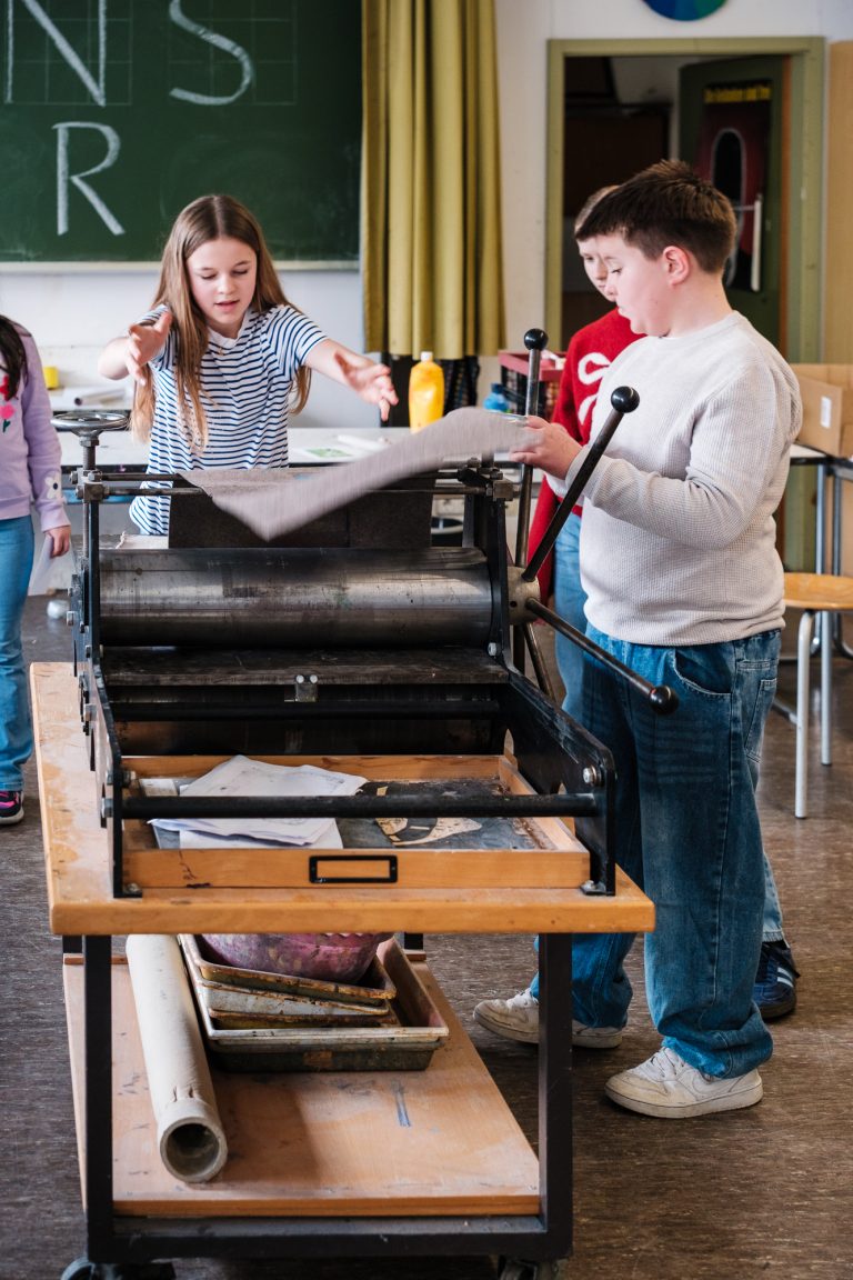 Schülerin und Schüler drucken ein Bild mit einer Druckerpresse