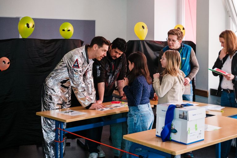 Lehrer im Astronautenkostum unterstützt Schülerinnen beim Lösen von Rechenaufgaben im Mathe-Escape-Room