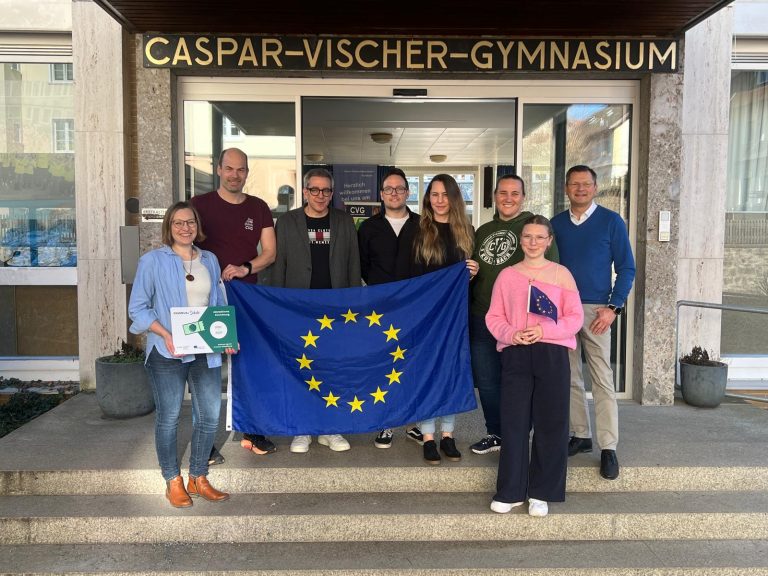 Lehrkräfte des Erasmus+ Teams, der Schulleiter und eine Schülersprecherin Mit EU-Flagge vor dem Haupteingang der Schule
