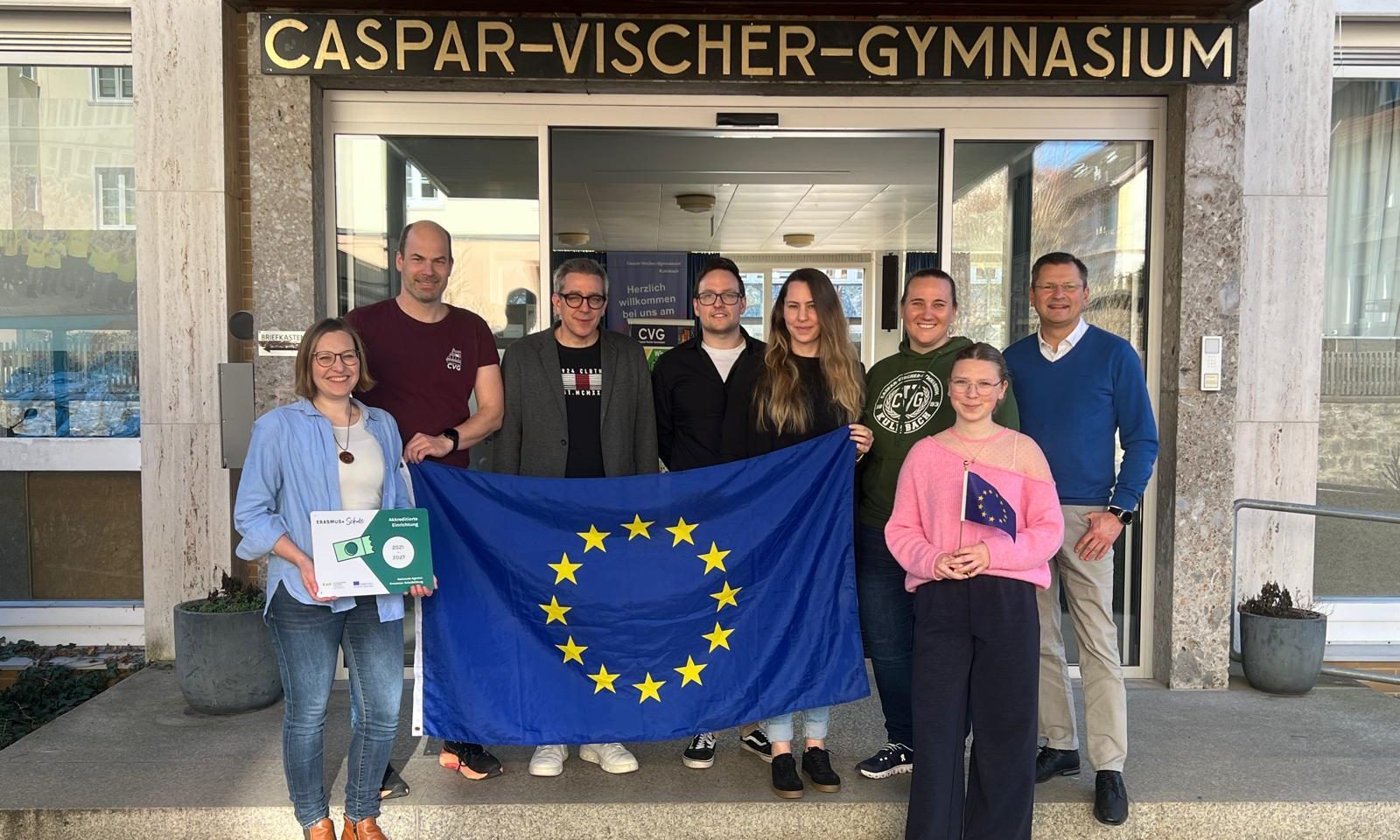 Lehrkräfte des Erasmus+ Teams, der Schulleiter und eine Schülersprecherin Mit EU-Flagge vor dem Haupteingang der Schule