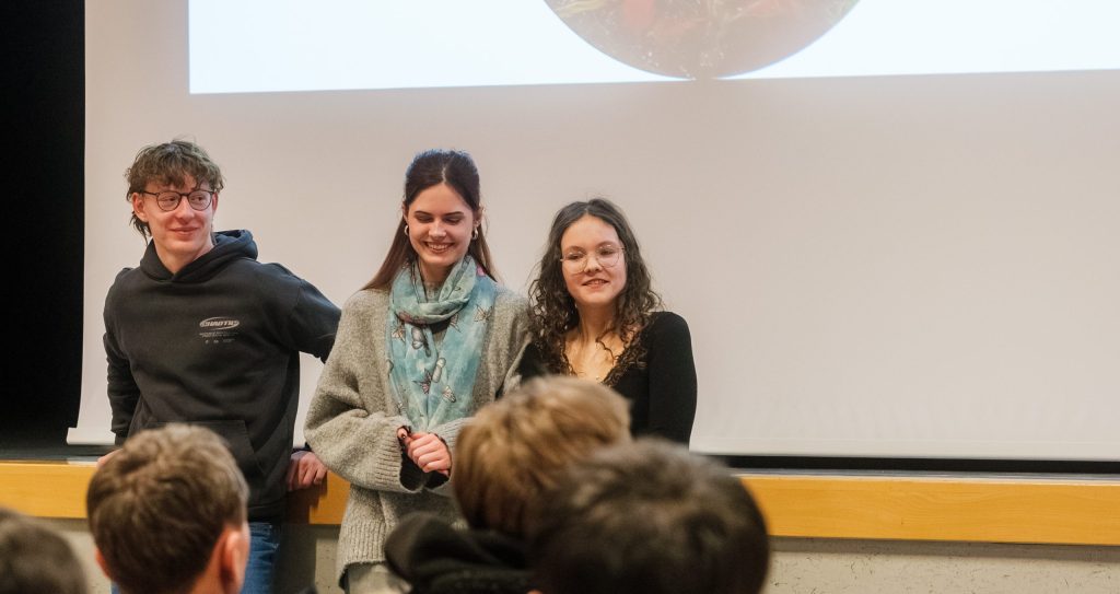 Drei Schülerinnen und Schüler stehen vor Publikum und berichten über die Einführungsklasse